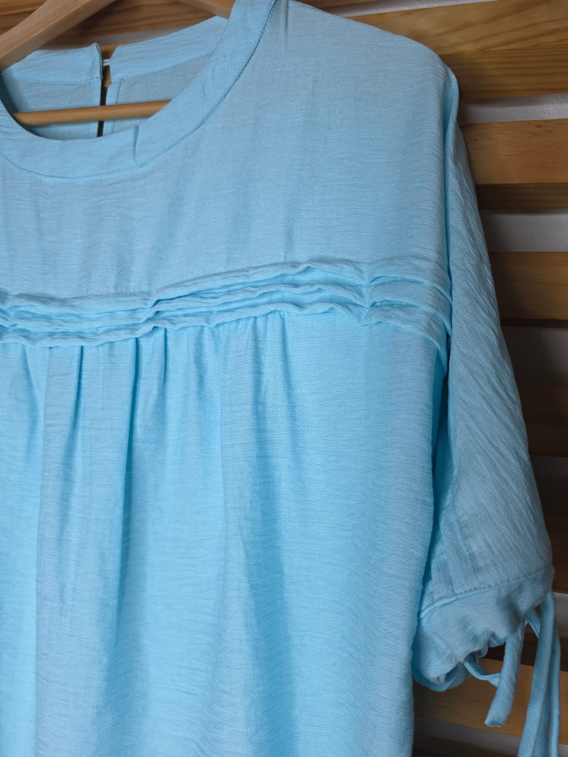 A light blue women's blouse with a round neckline, pleated details, short sleeves, and adjustable tie cuffs.