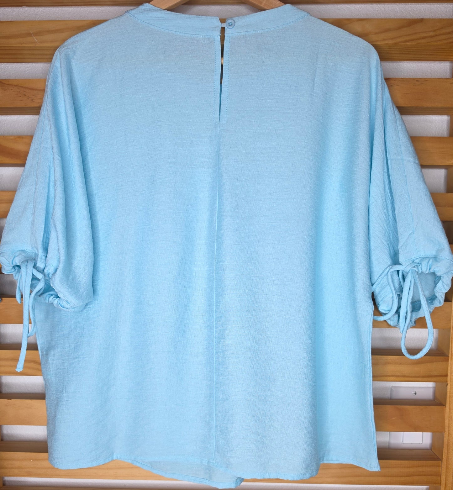 A light blue women's blouse with a round neckline, pleated details, short sleeves, and adjustable tie cuffs.