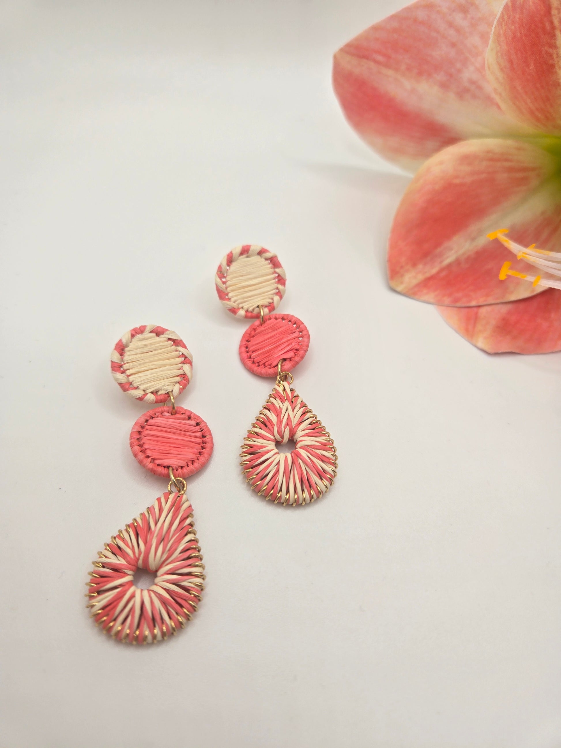A pair of handmade drop earrings with pink and cream striped woven details, displayed on a white surface next to a pink flower.