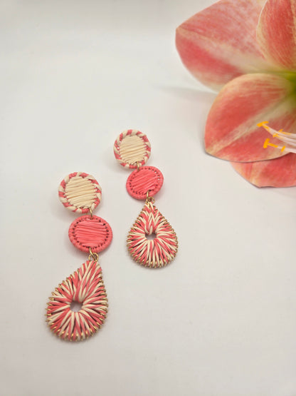 A pair of handmade drop earrings with pink and cream striped woven details, displayed on a white surface next to a pink flower.