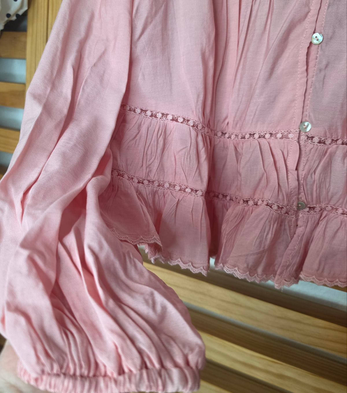 Women’s pink blouse with V-neckline, button front, lace-trimmed ruffles, delicate pin-tuck details, and long balloon sleeves with elastic cuffs.