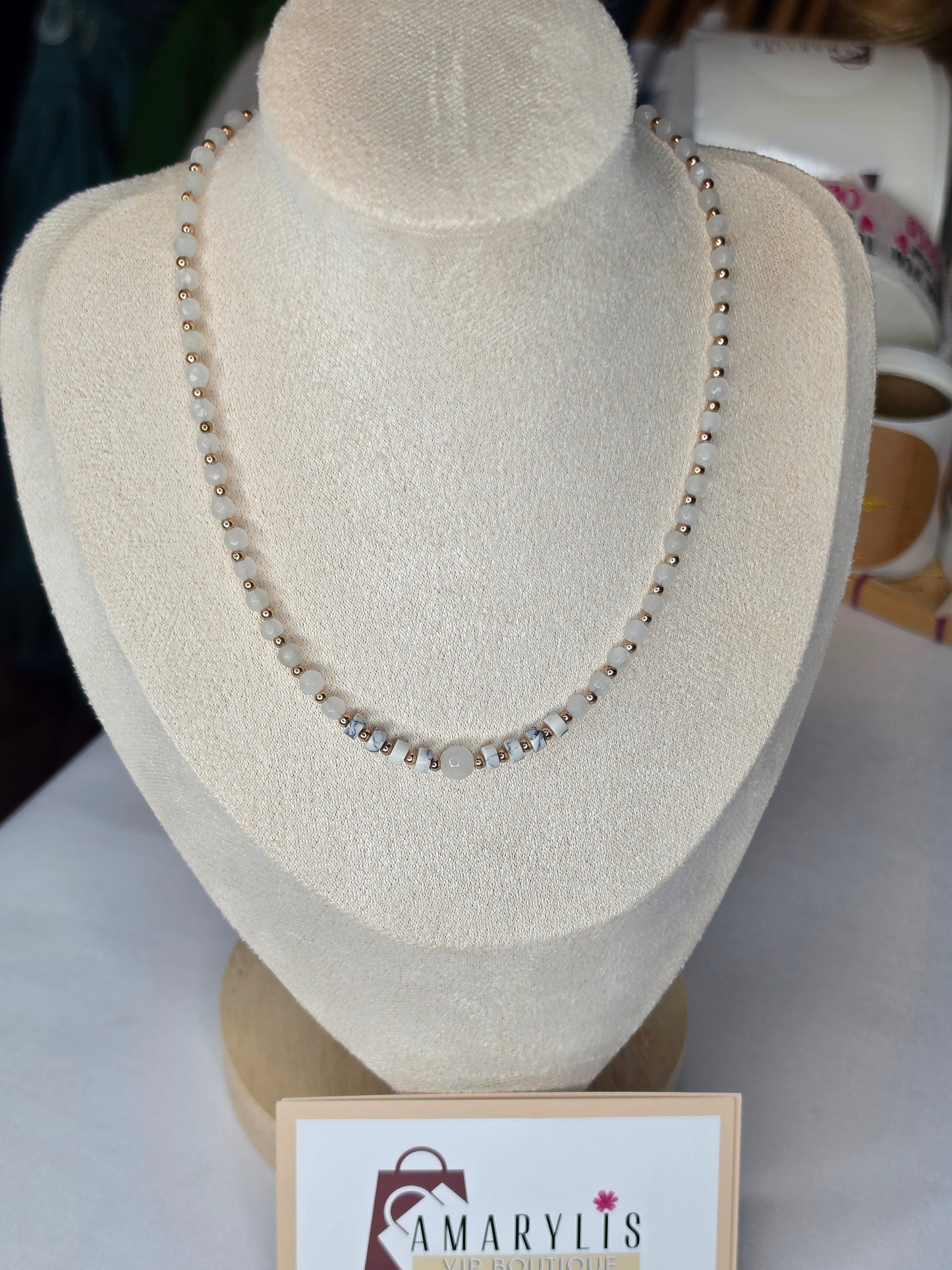 A beaded necklace displayed on a cream-colored jewelry bust. The necklace features small metallic beads with a central cluster of clear rhinestones and a larger central pearl-like bead.