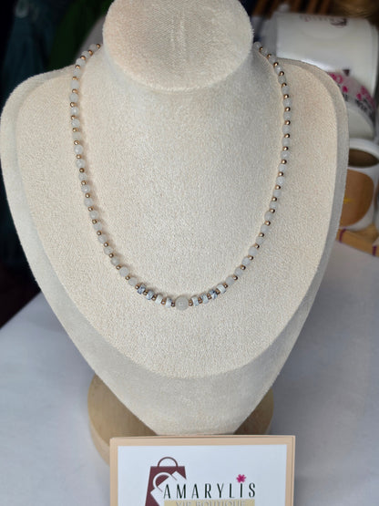 A beaded necklace displayed on a cream-colored jewelry bust. The necklace features small metallic beads with a central cluster of clear rhinestones and a larger central pearl-like bead.