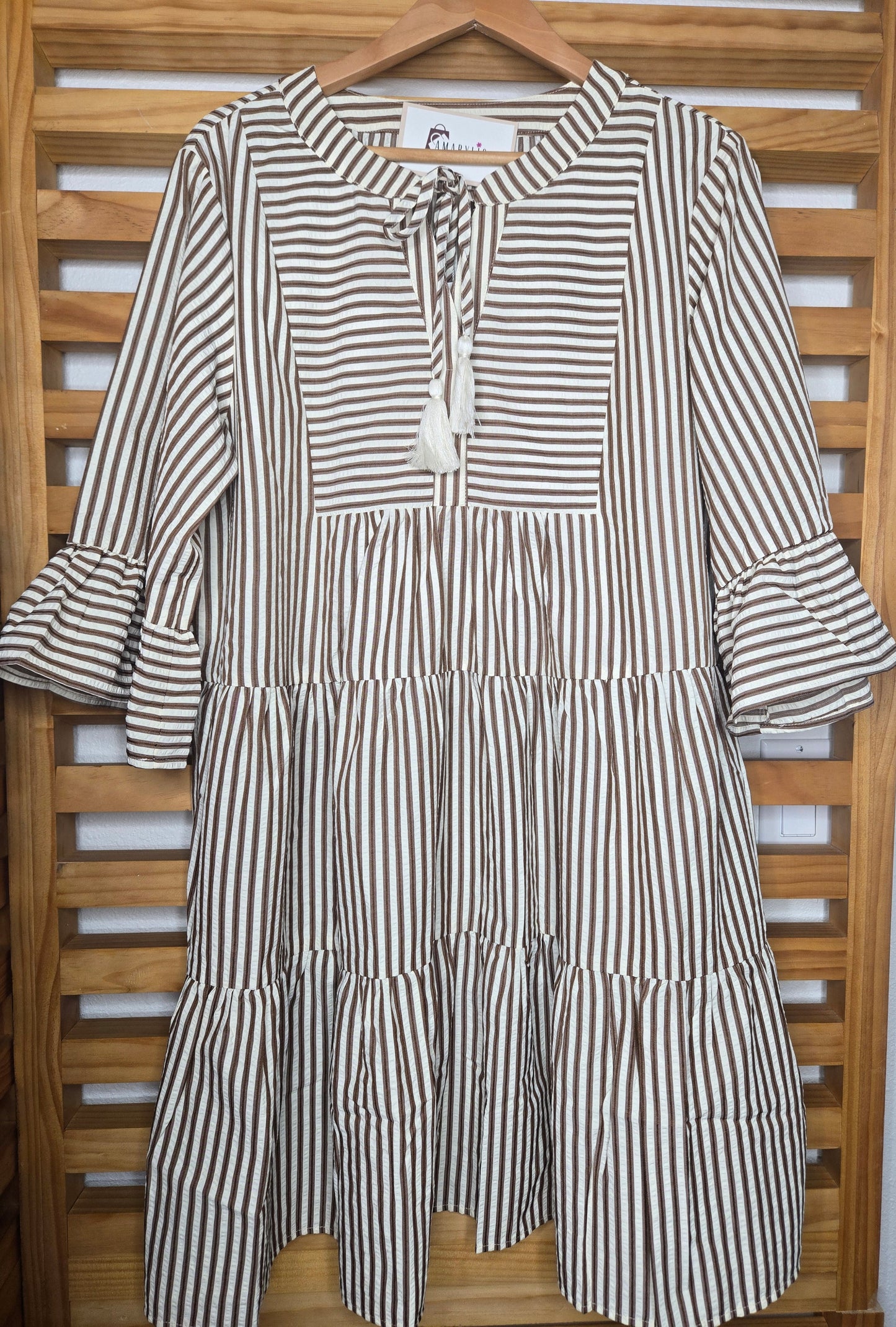 Lightweight cotton dress featuring classic color chocolate stripes, flowy bell sleeves, and a tassel tie neckline for a relaxed boho-inspired look. The loose silhouette offers comfort and movement, while the side pockets add everyday practicality. A charming and feminine piece that’s perfect for casual days or relaxed outings