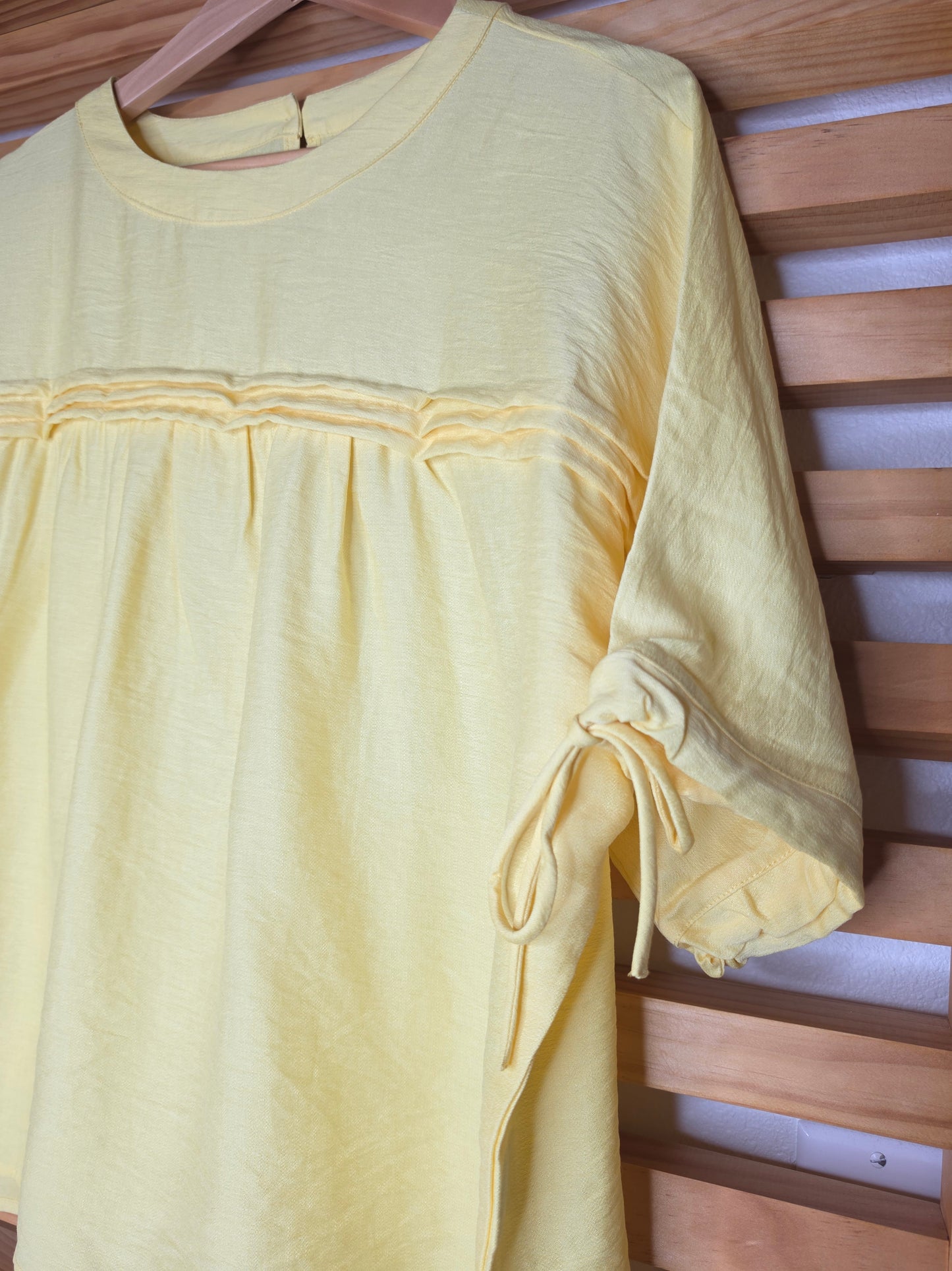 A yellow women's blouse with a round neckline, pleated details, short sleeves, and adjustable tie cuffs.