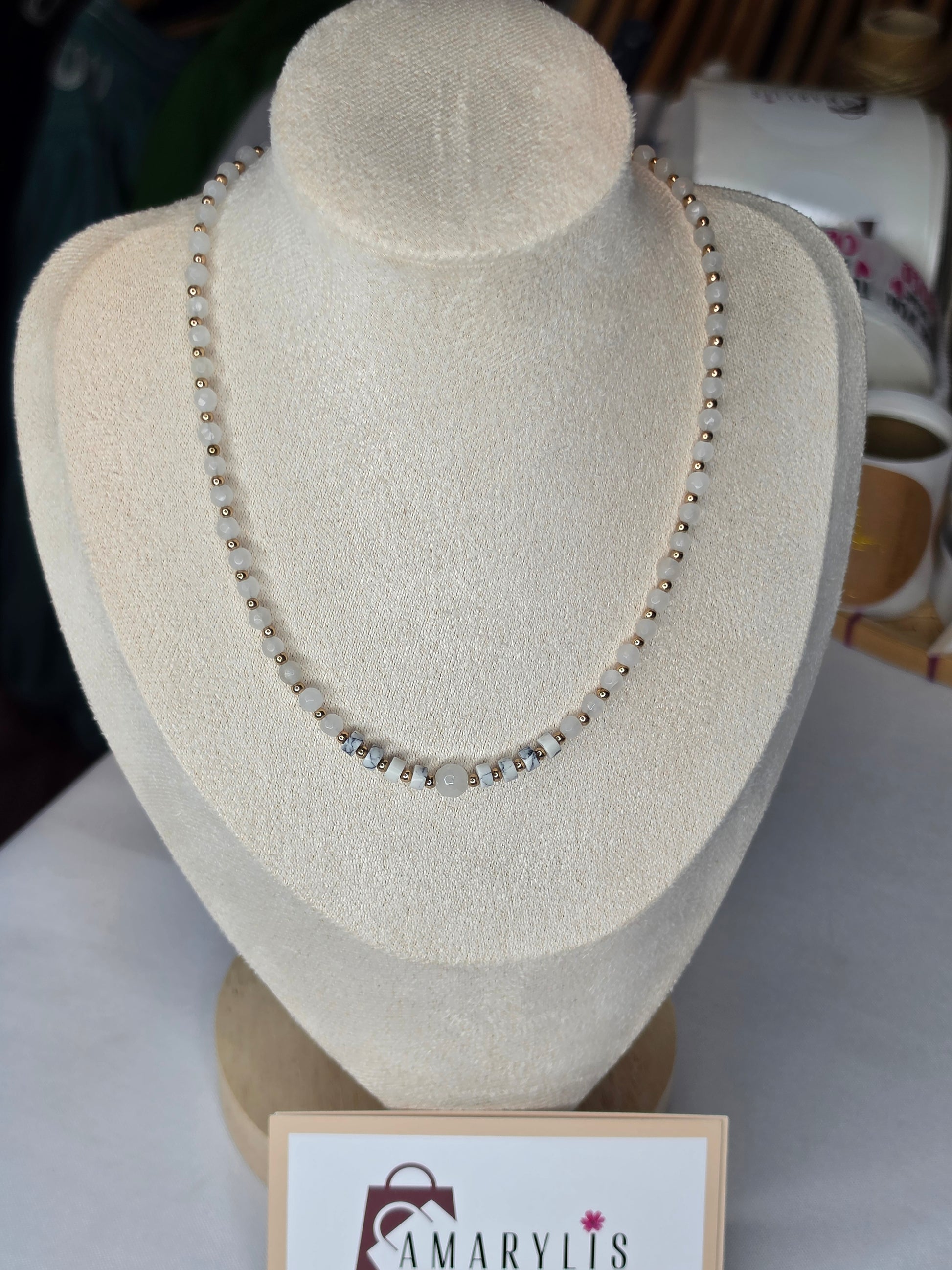 A beaded necklace displayed on a cream-colored jewelry bust. The necklace features small metallic beads with a central cluster of larger clear faceted beads.