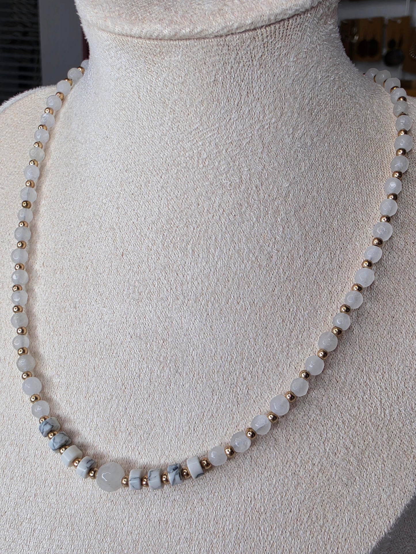 A beaded necklace displayed on a cream-colored fabric mannequin bust. The necklace features round, pale grayish-white beads separated by small metallic gold spacer beads, with a few larger polished pale blue-gray beads near the bottom left.