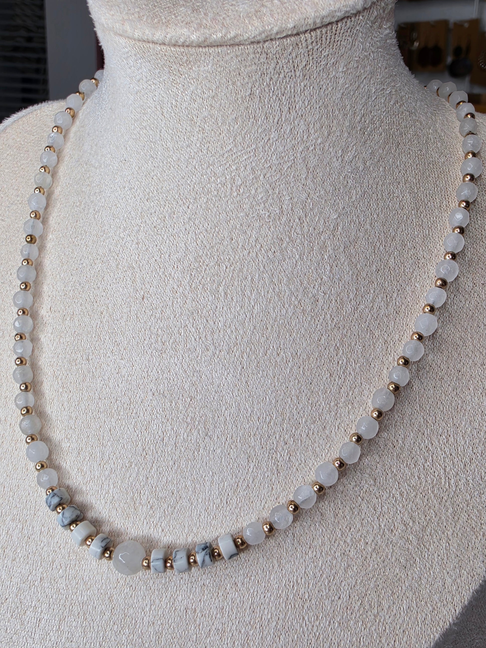 A beaded necklace displayed on a cream-colored fabric mannequin bust. The necklace features round, pale grayish-white beads separated by small metallic gold spacer beads, with a few larger polished pale blue-gray beads near the bottom left.