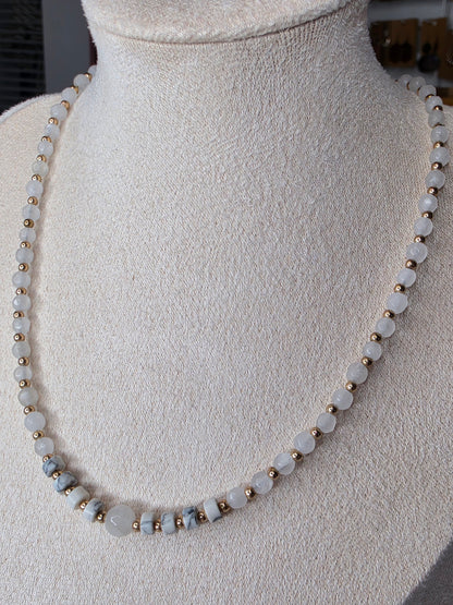 A beaded necklace displayed on a cream-colored fabric mannequin bust. The necklace features round, pale grayish-white beads separated by small metallic gold spacer beads, with a few larger polished pale blue-gray beads near the bottom left.