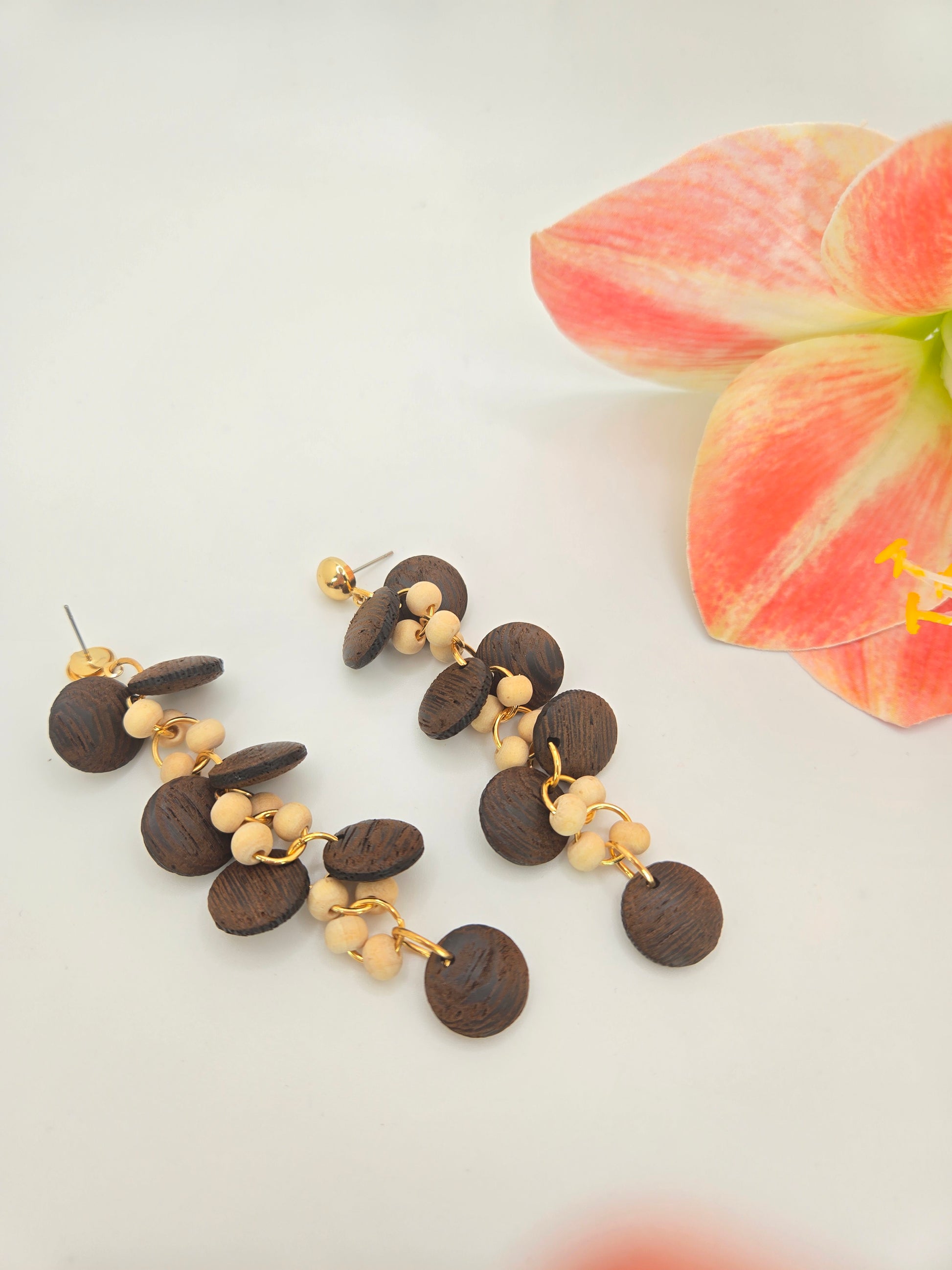A pair of dangling earrings displayed on a black T-stand. Each earring has gold-tone studs and hooks with layered dark brown wooden discs and small cream-colored beads.