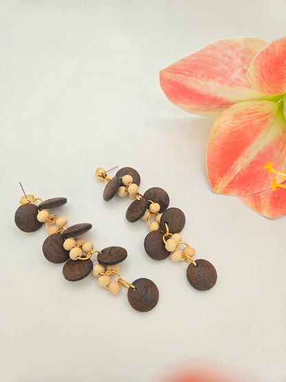 A pair of dangling earrings displayed on a black T-stand. Each earring has gold-tone studs and hooks with layered dark brown wooden discs and small cream-colored beads.