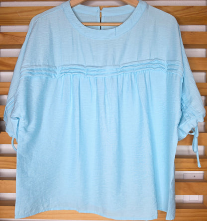 A light blue women's blouse with a round neckline, pleated details, short sleeves, and adjustable tie cuffs.